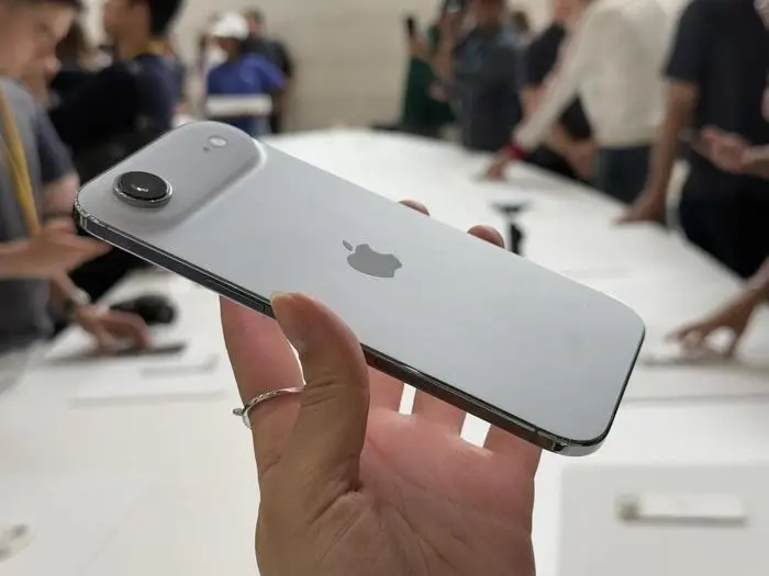 A hand holding a silver iPhone Air, displaying the Apple logo on the back and a camera module on the upper left. The device is shown in a bright, indoor setting with people in the background.